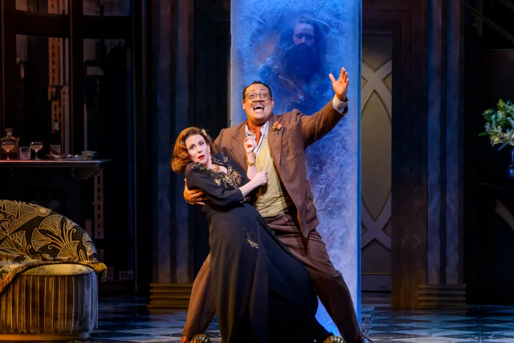 A man and woman in formal attire pose dramatically on stage in front of a frosted glass panel with a shadowy figure behind it.
