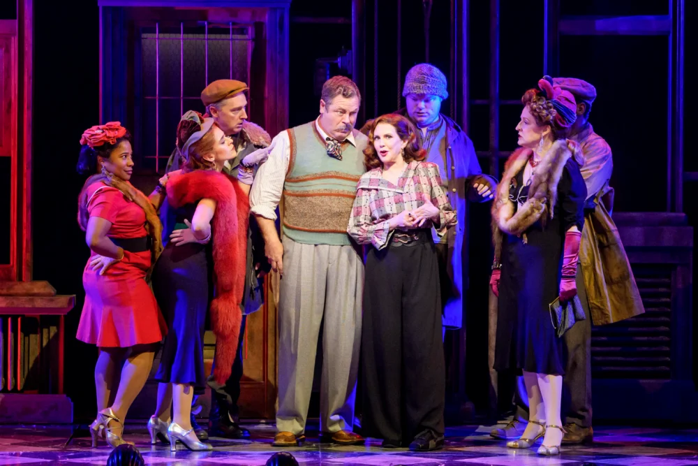 A group of actors in vintage costumes perform on stage, gathered in conversation under theatrical lighting and dramatic set pieces.