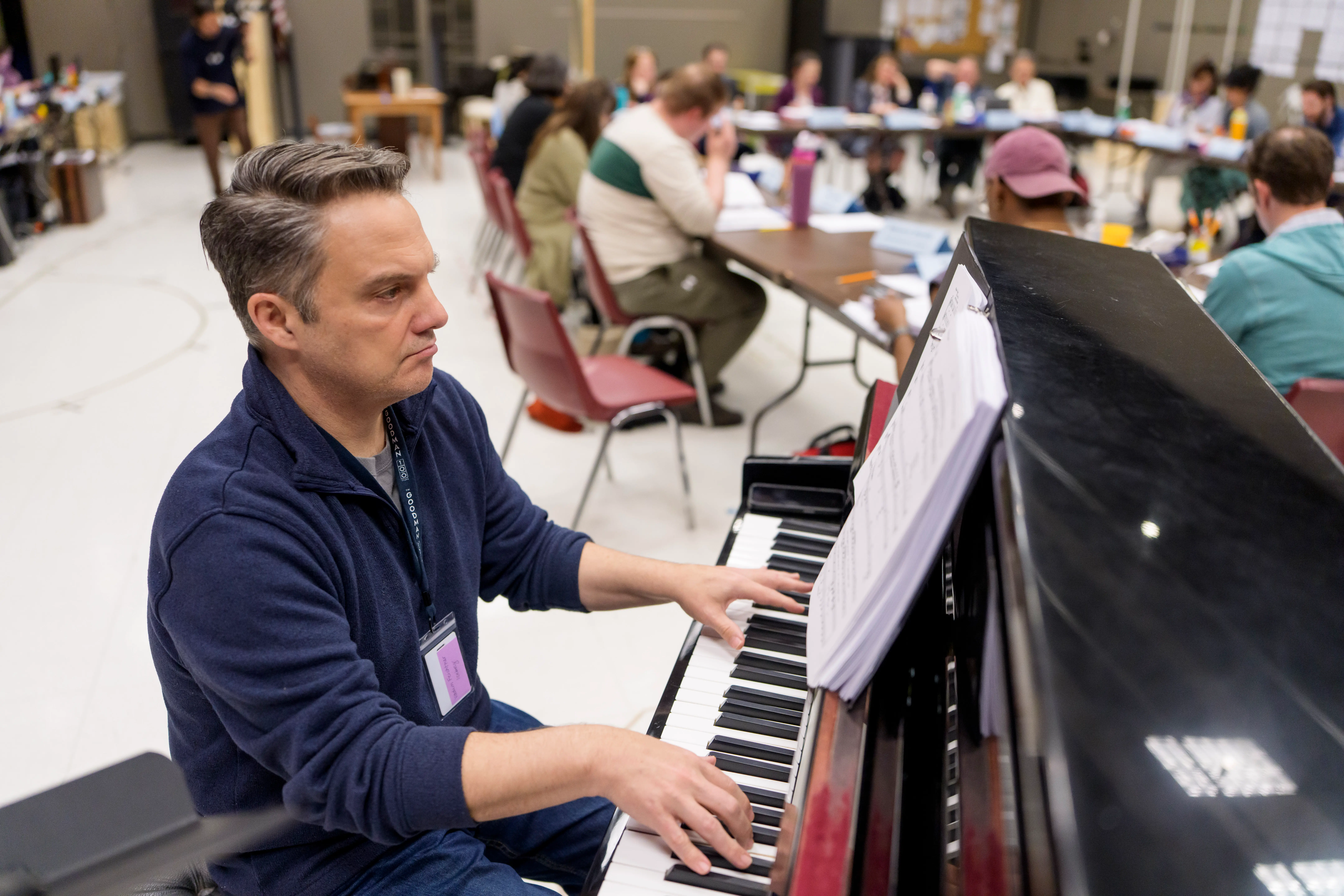 A man plays the piano while a group of people sit around tables in the background, appearing to participate in a workshop or rehearsal in a large room.