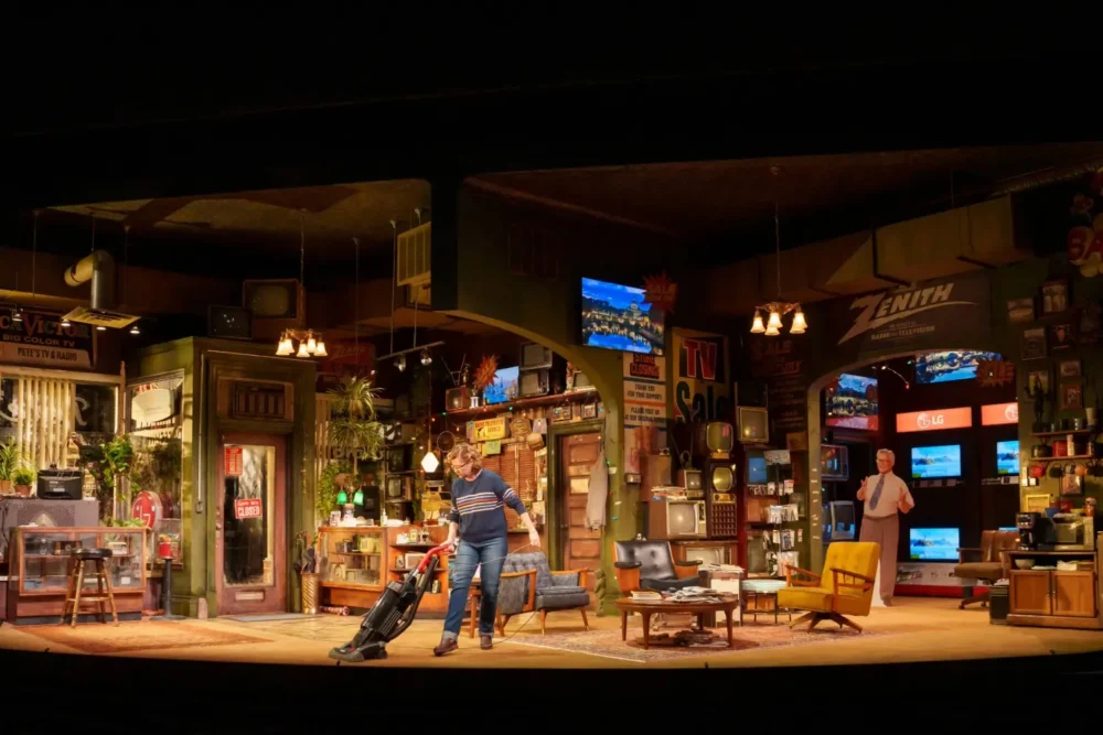 Stage set of a cluttered electronics store with two people inside, one vacuuming the floor and another standing near televisions and shelves filled with various items.