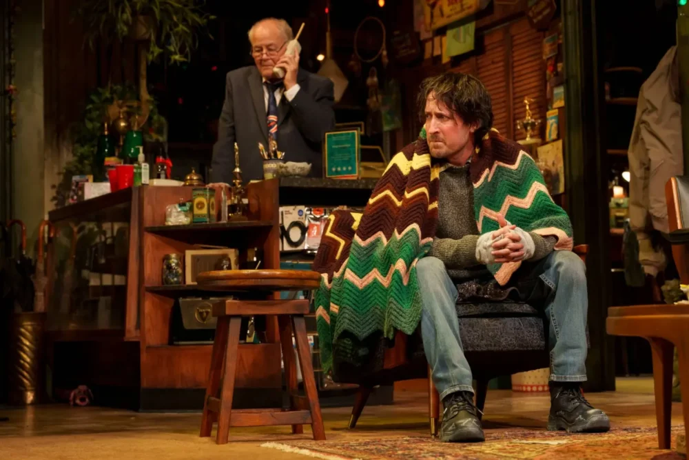 Two men are in a cluttered room; one sits with a cup and a blanket over his shoulders, while the other stands behind a counter, speaking on the phone.