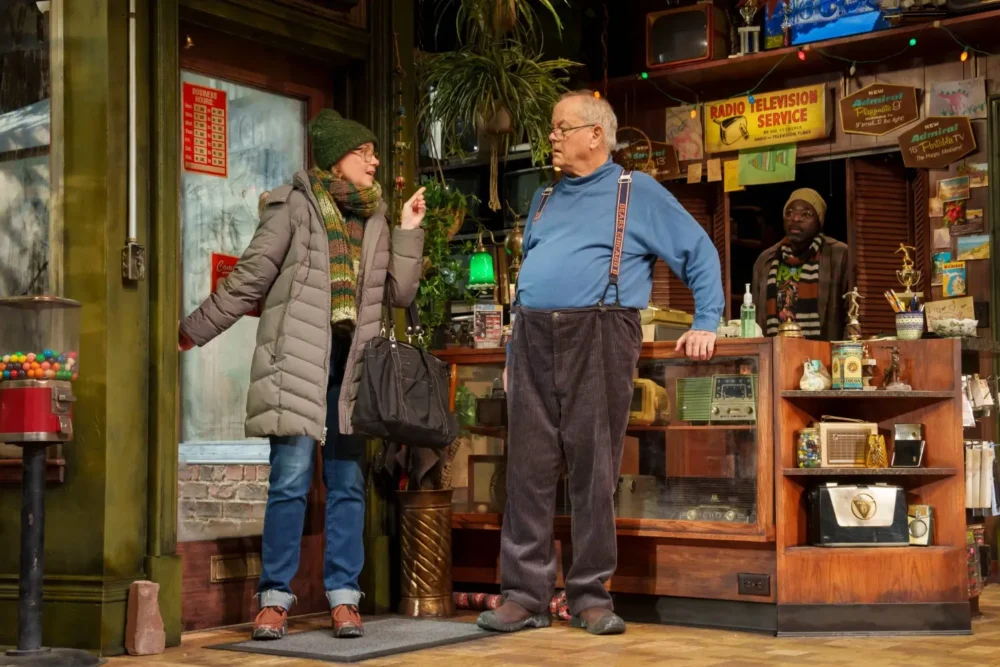 A woman in winter clothes speaks to an older man behind a counter in a cluttered vintage store, with another man standing in the background.