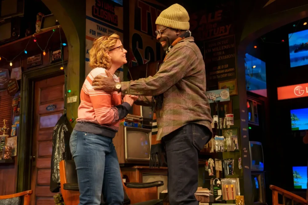 Two people stand facing each other and smiling on a cluttered, retro-themed stage set with various vintage items and colorful string lights.
