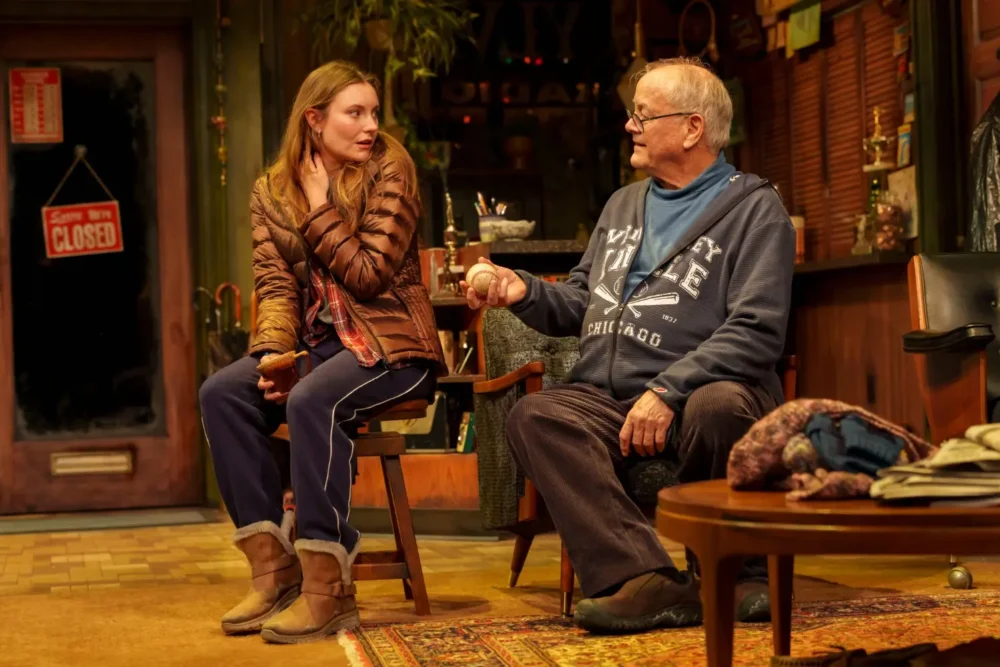 A young woman in casual clothes sits next to an older man holding a cup in a cozy, cluttered room with warm lighting and a "Closed" sign on the door in the background.