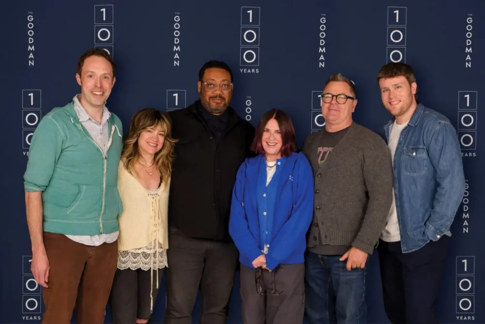 Six people stand in a row, smiling in front of a blue Goodman Theatre 100 Years backdrop.