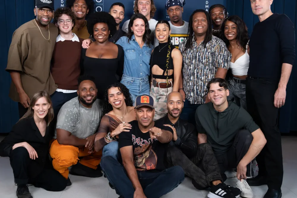 A group of eighteen people pose together indoors, arranged in three rows, smiling at the camera against a dark blue background.