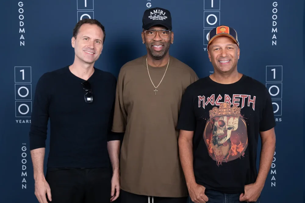 Three men stand side by side in front of a blue backdrop with "The Goodman 100 Years" printed on it. They are smiling and posing for the photo.