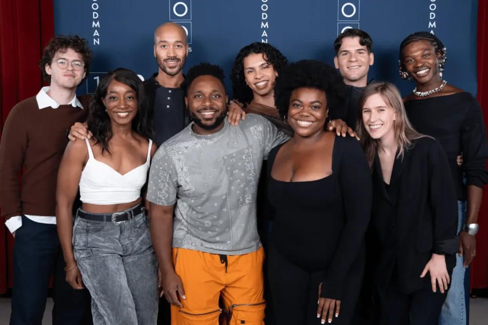 A group of nine people stand together, smiling for a photo in front of a blue backdrop with "Goodman" written on it.