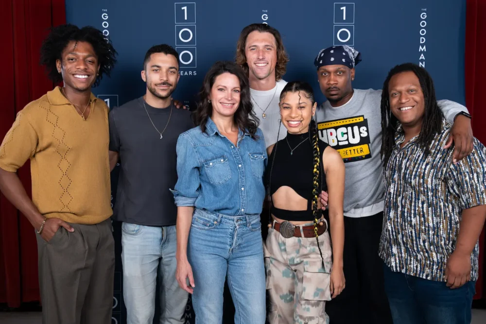 Seven people stand together in front of a blue backdrop with "100 Years" and "The Goodman" logos, posing and smiling for a group photo.