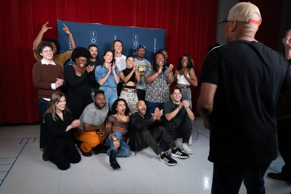 A group of people pose and smile together in front of a blue backdrop, while a person in a black shirt and cap stands facing them in the foreground.