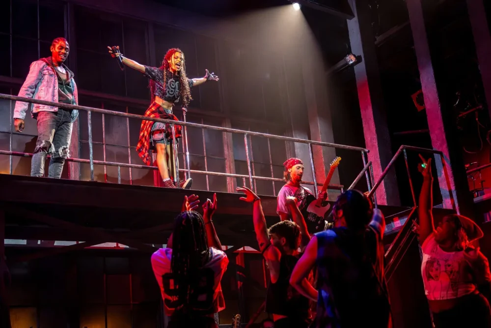 Performers on a stage, with one person standing with arms outstretched on an upper level, others playing guitar and people in the audience reaching up toward them.