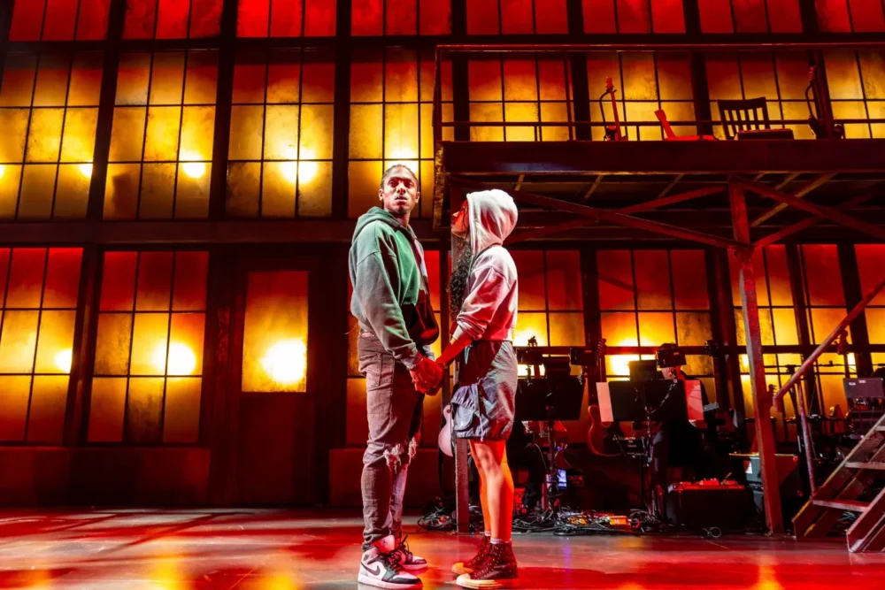 Two people stand holding hands onstage, with a dramatic red and orange lit background and musicians seated below a balcony set.