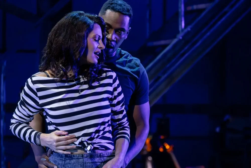 A man stands closely behind a woman in a black-and-white striped shirt, holding her around the waist, both appearing engaged in an intimate moment on stage under blue lighting.