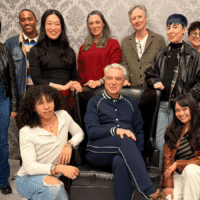 A group of twelve people posing together indoors, with one person seated in a black armchair and the others standing or sitting around them against a patterned wallpaper background.