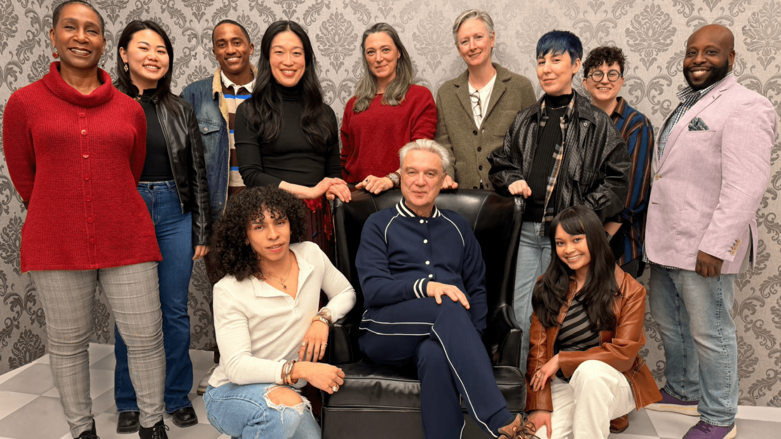 A group of twelve people posing together indoors, with one person seated in a black armchair and the others standing or sitting around them against a patterned wallpaper background.