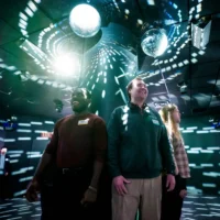 Three people stand together in a dark room with disco balls and patterned light projections on the walls and ceiling.