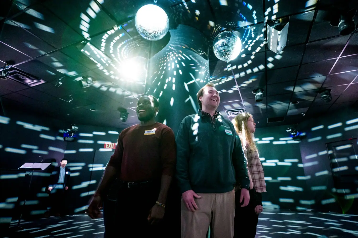 Three people stand together in a dark room with disco balls and patterned light projections on the walls and ceiling.