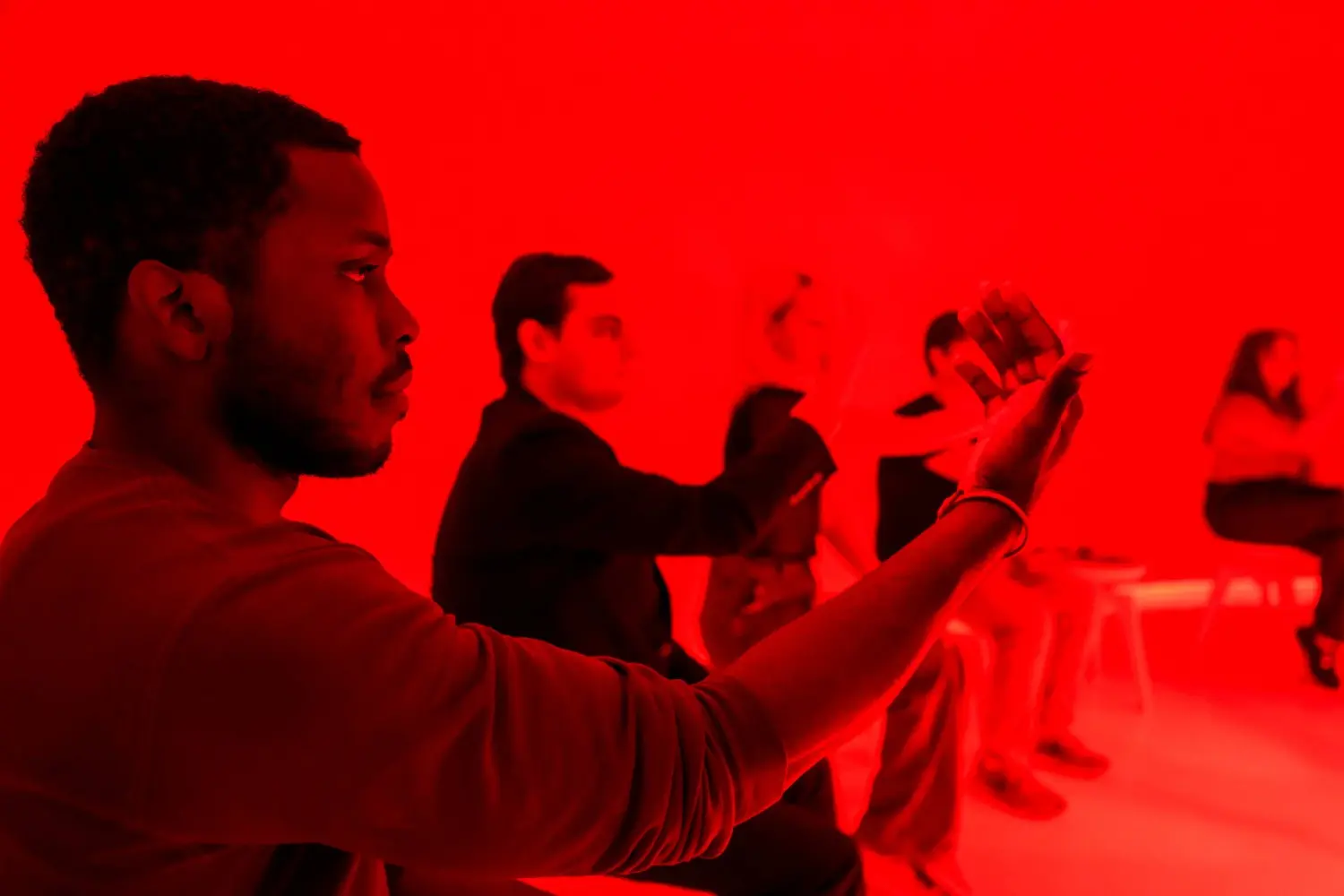 Several people sit on chairs in a room lit with intense red light; one person in the foreground holds up a phone, possibly taking a photo or video.