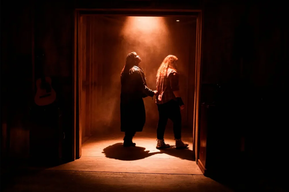 Two people walk through a dimly lit doorway, with one leading the other by the wrist. Light from above casts shadows on the wooden floor. A guitar is visible on the left.