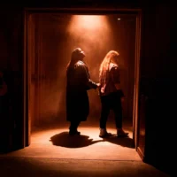 Two people walk through a dimly lit doorway, with one leading the other by the wrist. Light from above casts shadows on the wooden floor. A guitar is visible on the left.