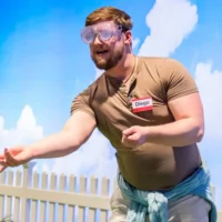 A man wearing safety goggles and a name tag labeled "Diego" throws an object, with a white fence and blue sky backdrop behind him.