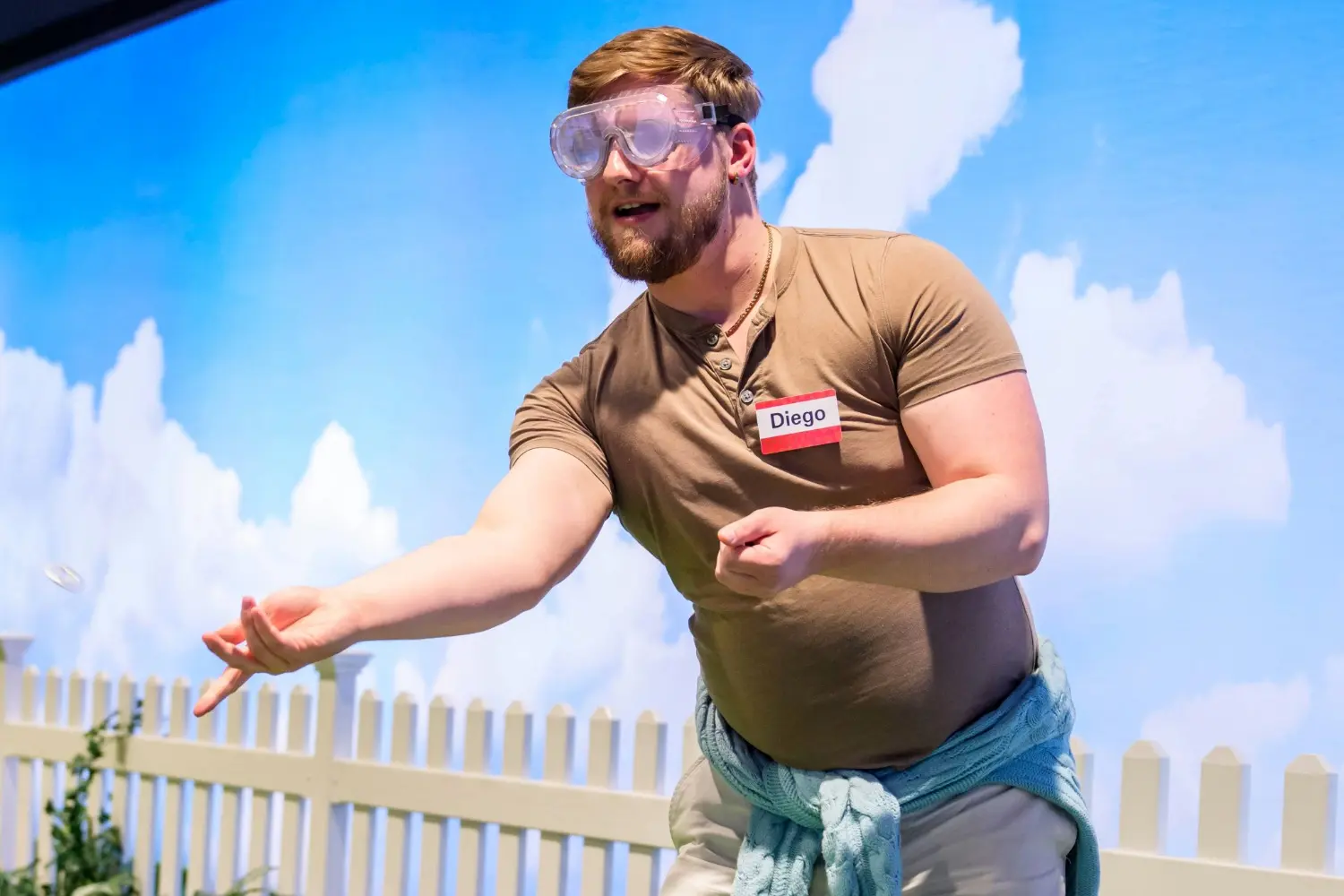 A man wearing safety goggles and a name tag labeled "Diego" throws an object, with a white fence and blue sky backdrop behind him.