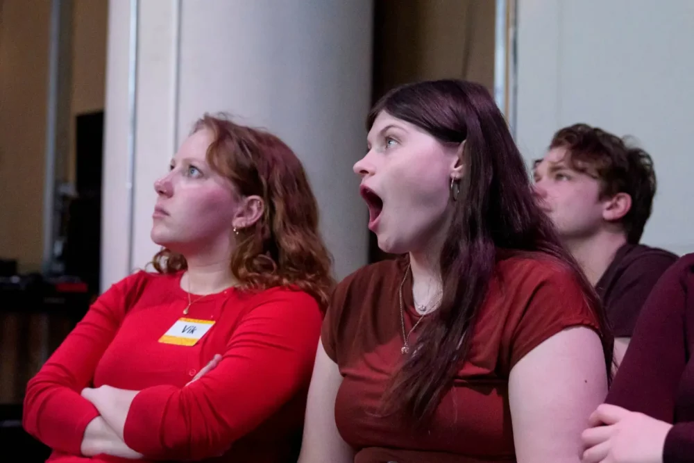 Three people sitting indoors; the person in the middle appears surprised or yawning, while the others look ahead with neutral expressions.