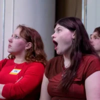 Three people sitting indoors; the person in the middle appears surprised or yawning, while the others look ahead with neutral expressions.