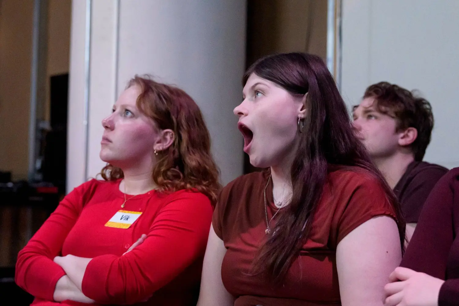 Three people sitting indoors; the person in the middle appears surprised or yawning, while the others look ahead with neutral expressions.