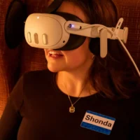 A person wearing a VR headset sits in a chair, with a visible name tag that reads "Shonda" on their shirt.