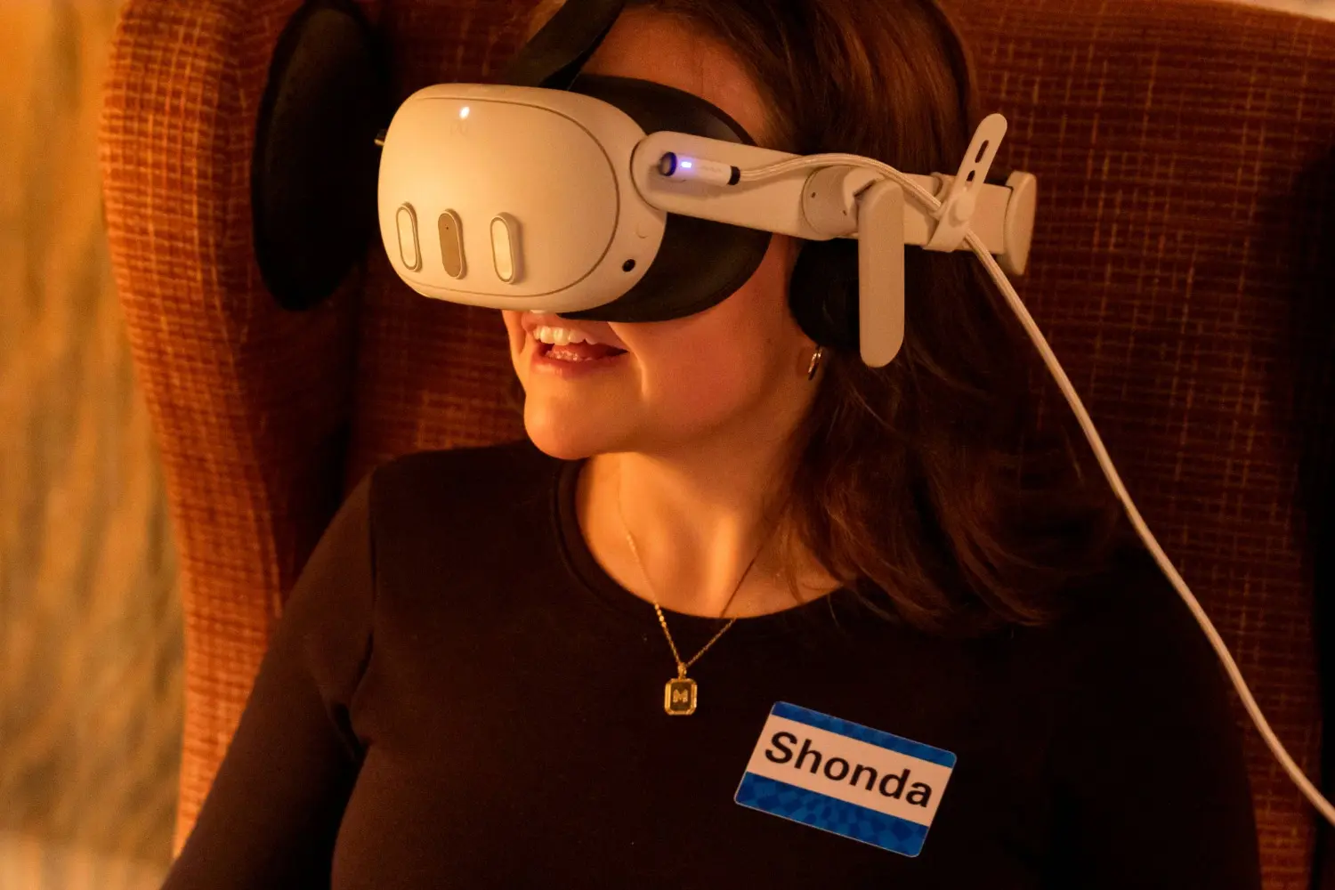 A person wearing a VR headset sits in a chair, with a visible name tag that reads "Shonda" on their shirt.