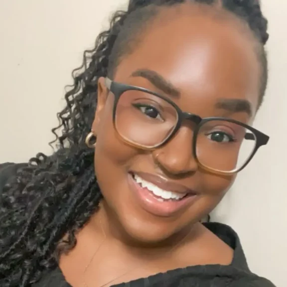 A woman with curly hair, glasses, and a black top smiles at the camera in a close-up portrait against a neutral background.