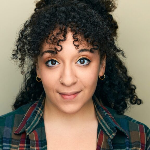 A person with curly hair and hoop earrings looks at the camera, wearing a plaid shirt and a nose ring, against a plain beige background.