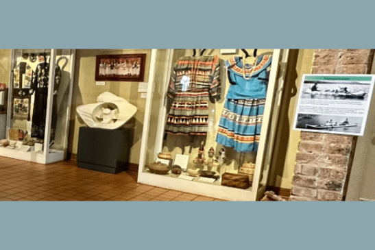 A museum display of Indigenous artwork and clothing, featuring an informational sign on the wall.
