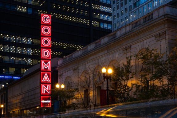 A vertical neon sign reading "GOODMAN" is illuminated on the side of a building at night in an urban area, glowing brightly as city lights hint at the Sweetest Season unfolding among the office buildings in the background.