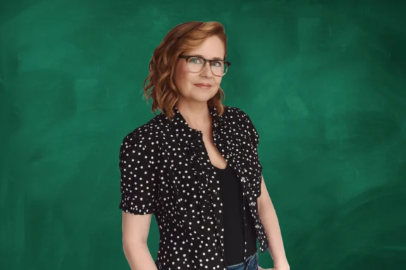 A woman with shoulder-length auburn hair, glasses, and a black polka dot shirt stands in front of a green chalkboard background.