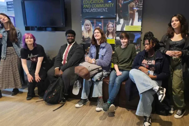 A group of six young people sit and stand indoors, smiling. Behind them is a poster reading "Education and Community Engagement" with photos of students.