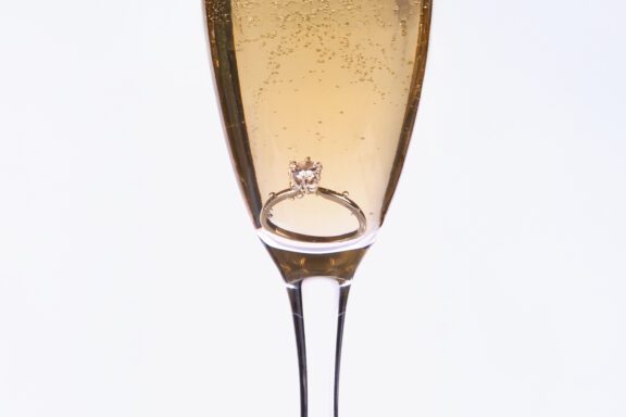 A diamond engagement ring sits at the bottom of a champagne flute filled with sparkling wine.