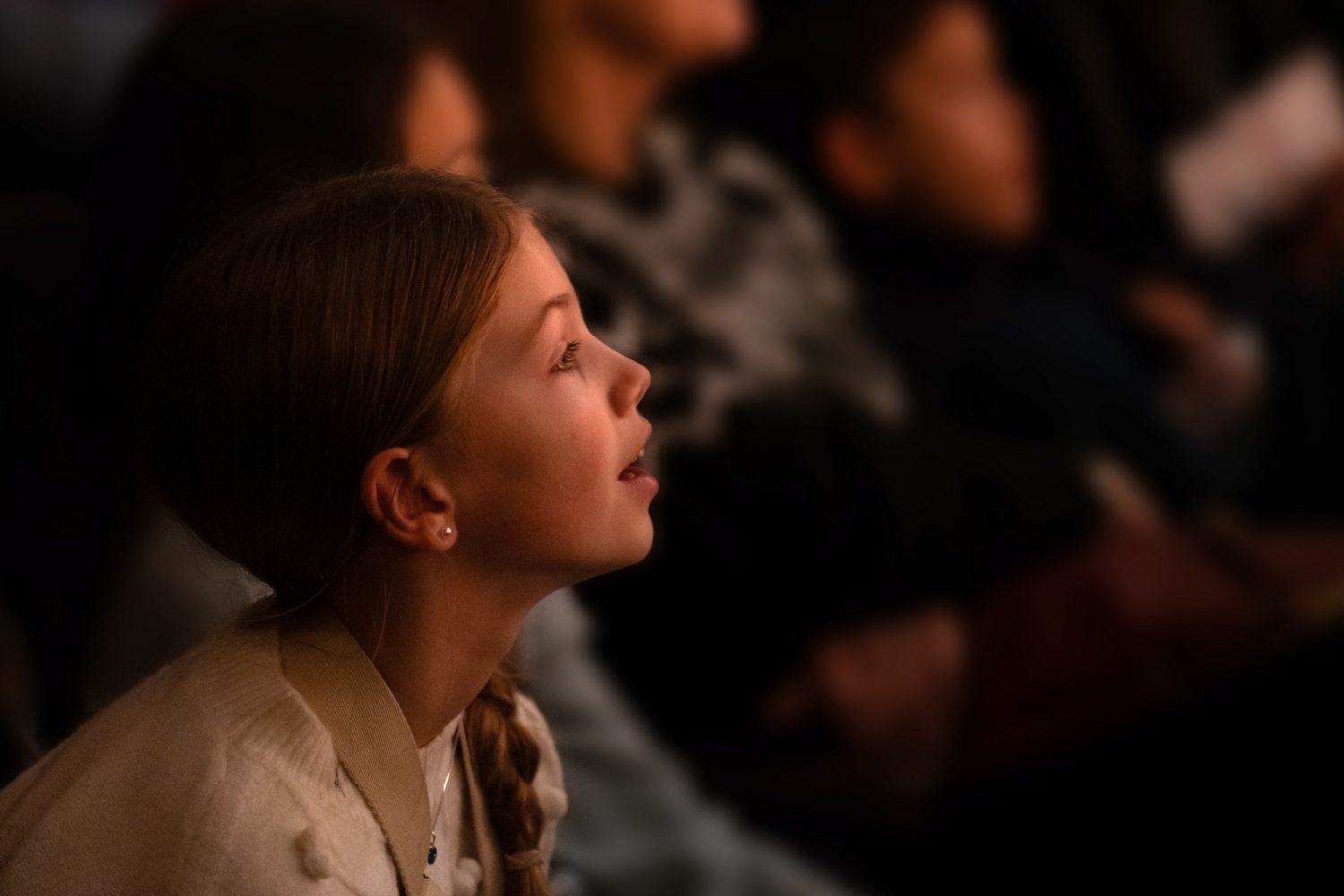 A young girl with braided hair sits in a crowd, looking ahead with interest and focus. Other people are seated nearby, slightly out of focus.