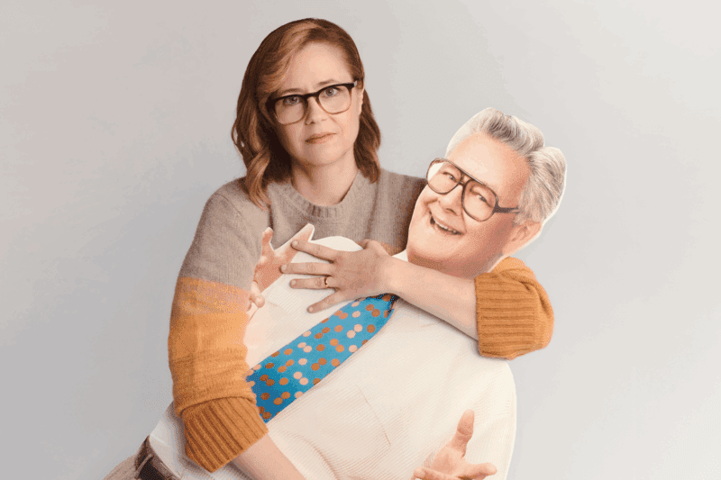 A woman with glasses holds a life-size cardboard cutout of a smiling older man wearing glasses, a white shirt, and a blue polka dot tie.