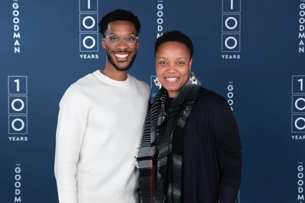 Two people smiling in front of a blue backdrop featuring "100 Years" and "The Goodman" logos.