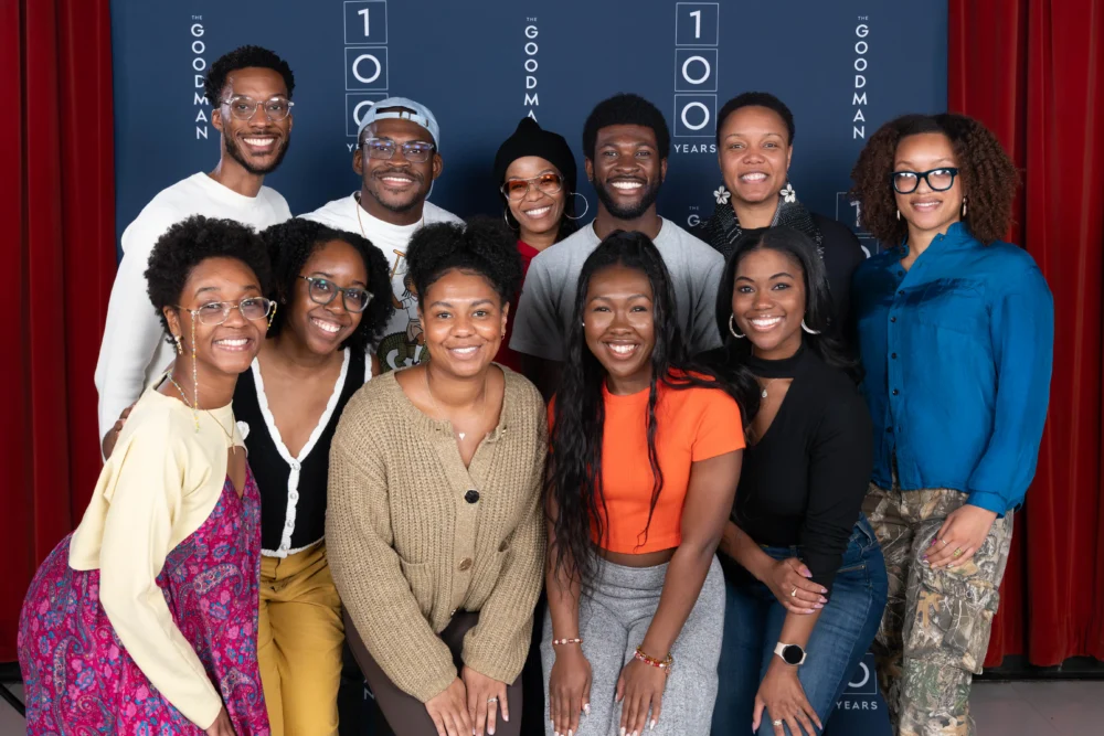 A group of twelve people stand and smile together in front of a backdrop with "GOODMAN 100 YEARS" text.