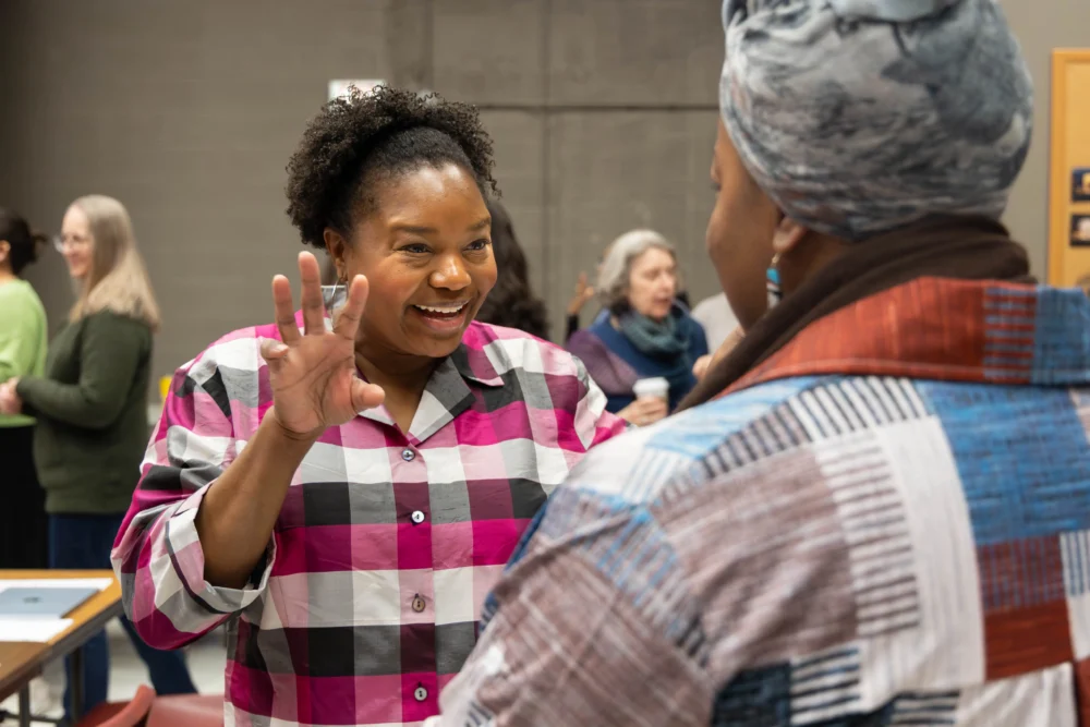 A woman in a pink plaid shirt gestures while speaking to another woman in a patterned headscarf at an indoor social gathering.
