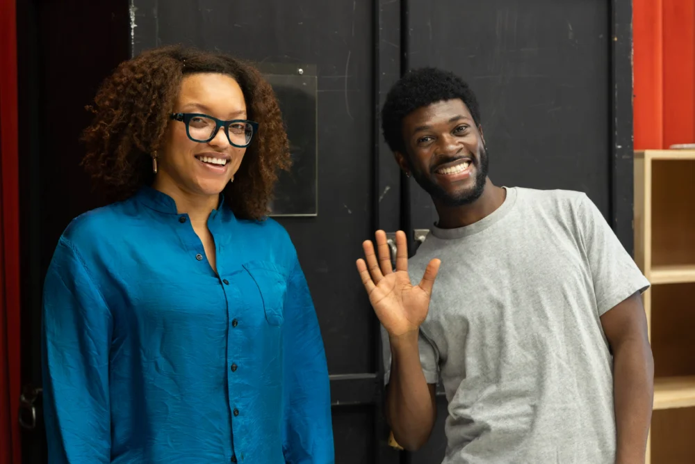 Two people stand indoors; one is smiling and waving, the other is wearing glasses and a blue shirt, both facing the camera against a black background.