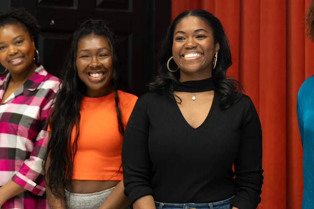 Three women stand indoors, smiling at the camera. The background features a dark door and red curtains.