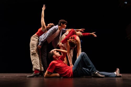 Five dancers pose on stage, four standing in a dramatic formation while one lies on the floor in front, all dressed in casual clothing against a dark background.