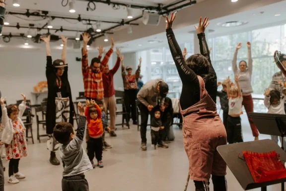 A group of adults and children stand indoors with arms raised, participating in a group activity or class.