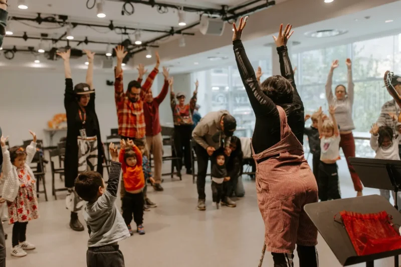 A group of adults and children stand indoors with arms raised, participating in a group activity or class.