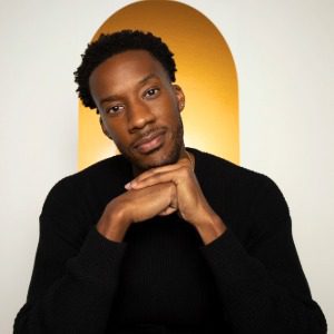 A man in a black sweater sits with his hands folded under his chin in front of a yellow arched background.