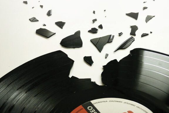 A black vinyl record lies broken, with several jagged pieces scattered around it on a white surface.